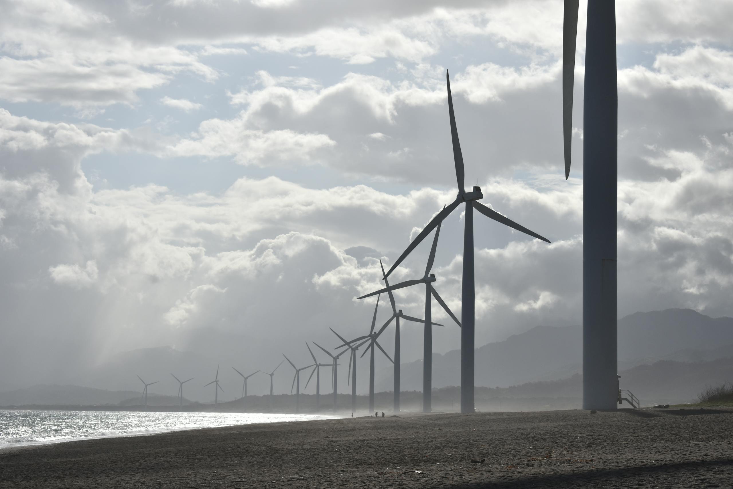 A row of wind turbines along a cloudy beach shoreline in Vigan City, Philippines.